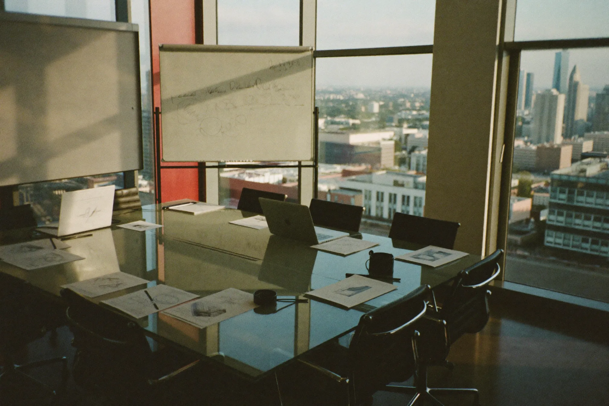 A modern consulting meeting room in Sandton with a glass table, laptops, printed strategy documents and a whiteboard, city skyline through the windows